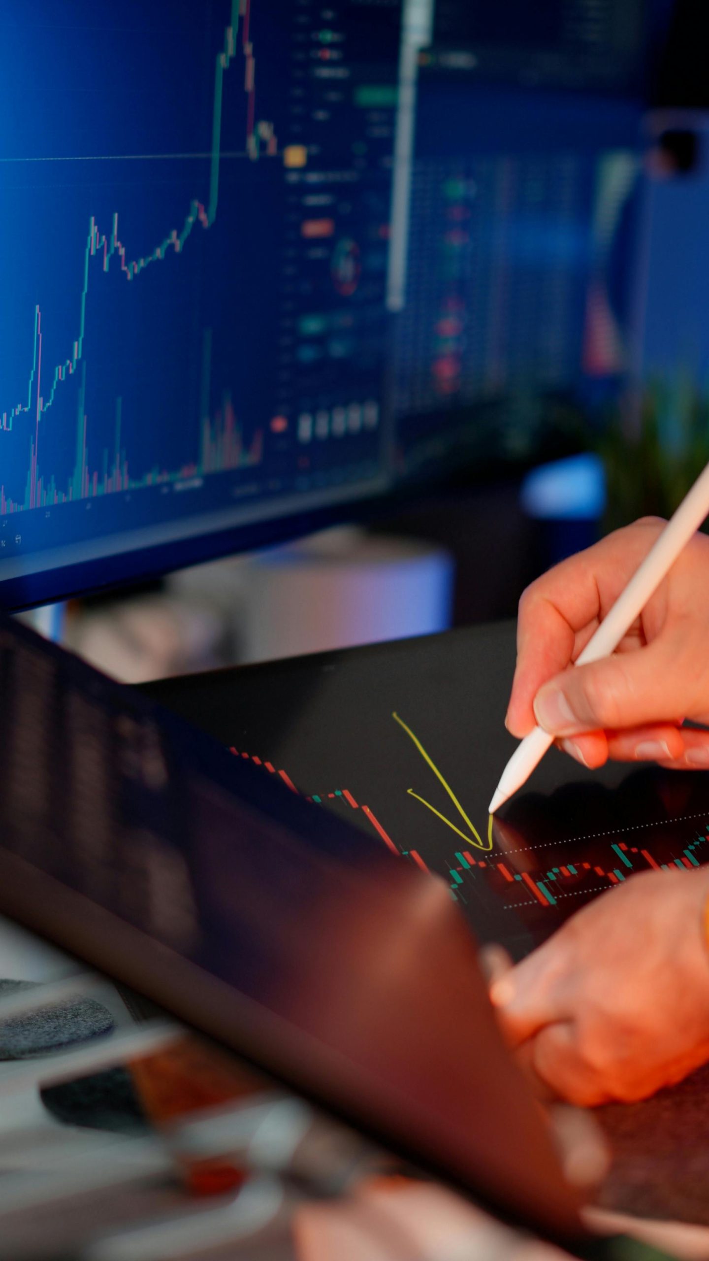 Person using a stylus on a tablet for stock market analysis, with charts on a monitor.